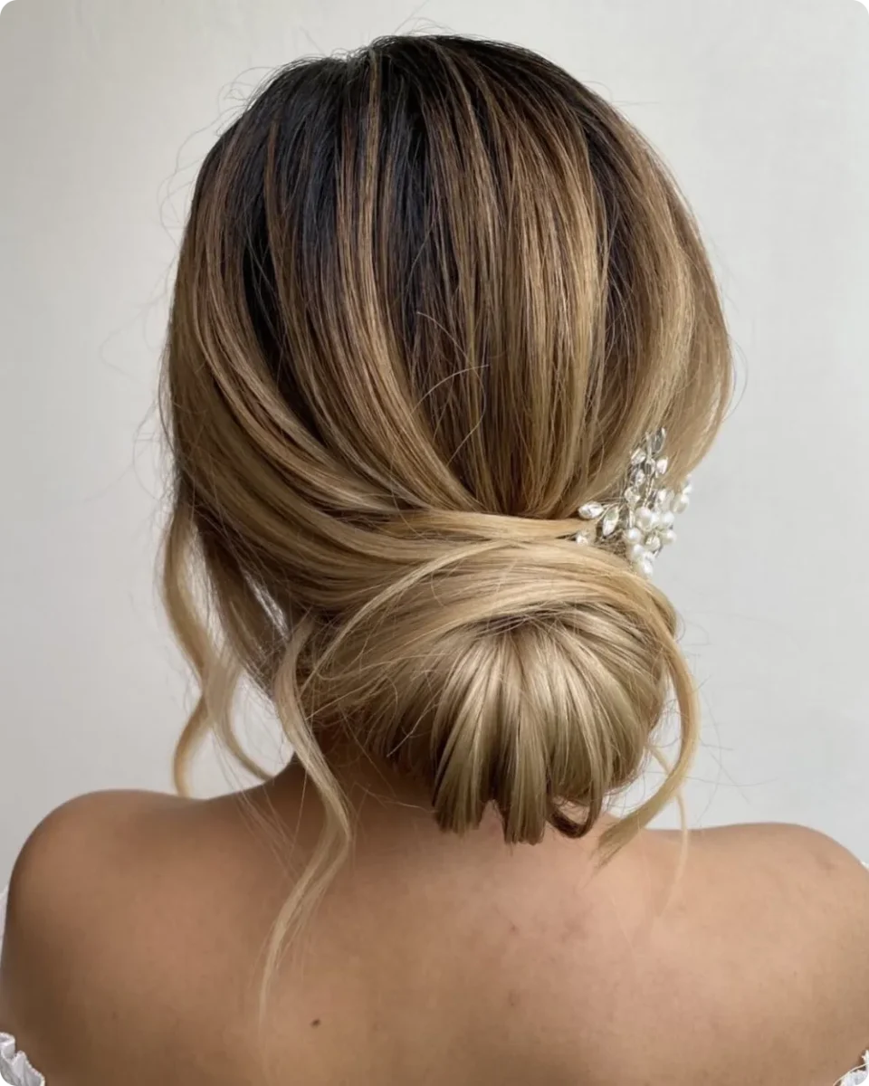 Low bun on balayage hair with dark roots and blonde ends, finished with a crystal and pearl comb on one side