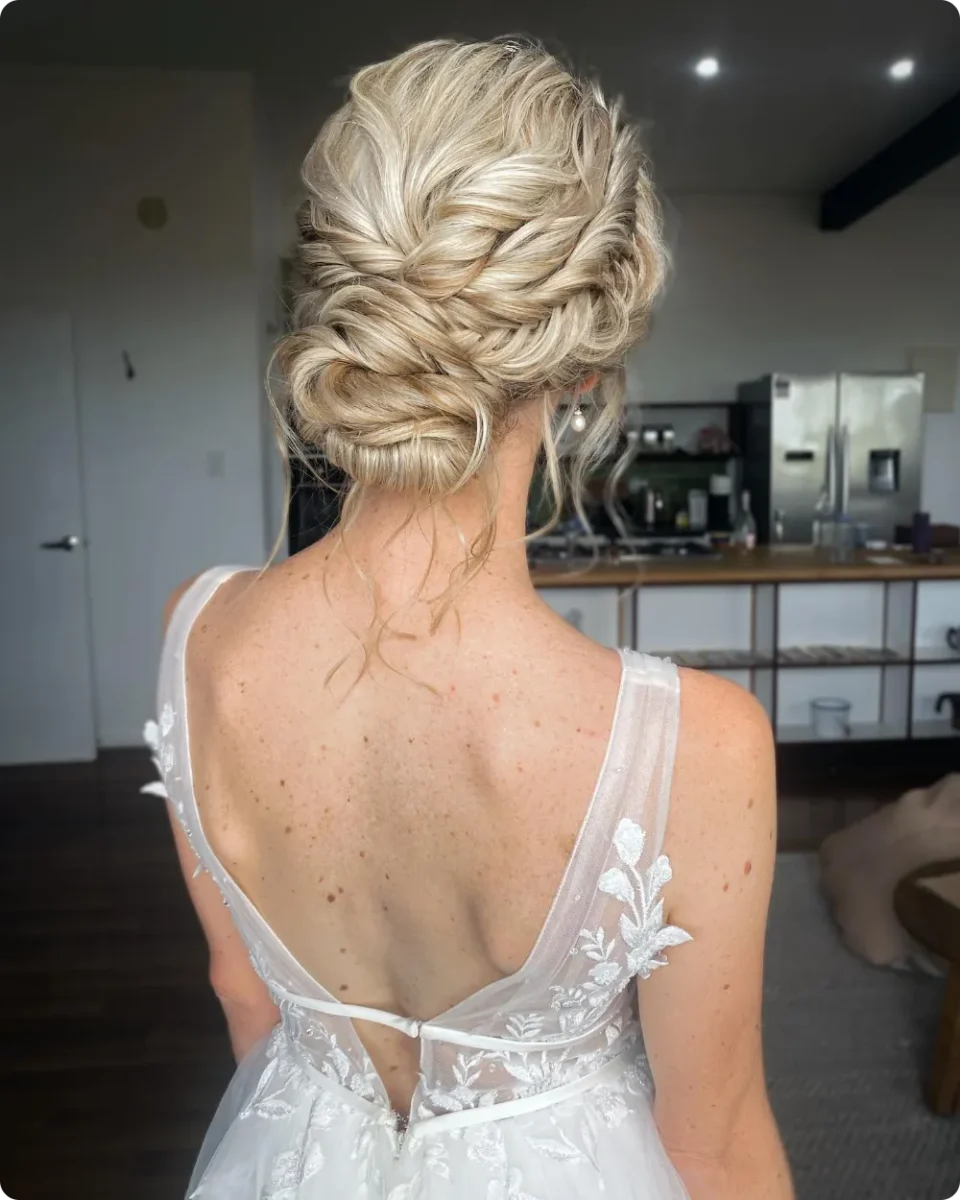 Braided boho bridal bun on light blonde hair with loose curls at the neck, worn with a low back lace wedding gown