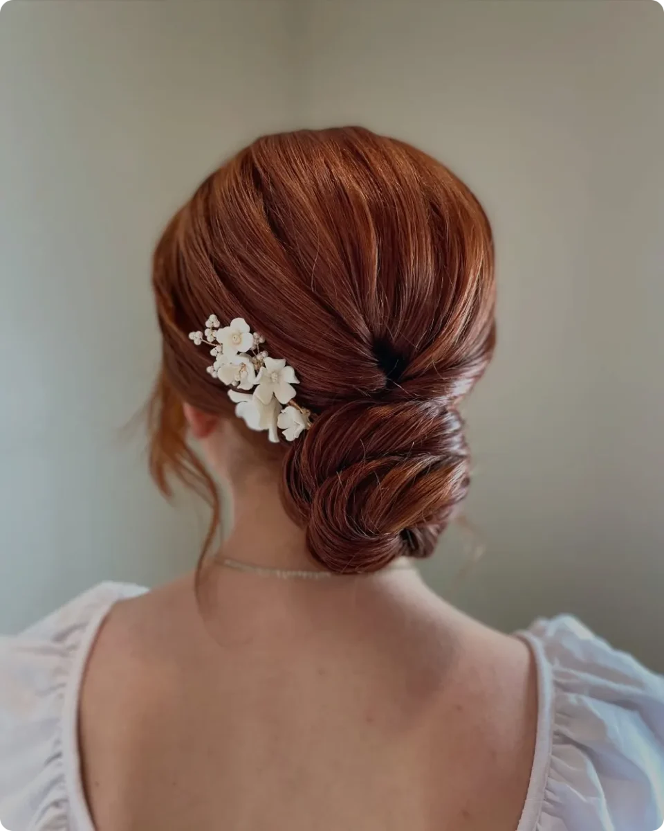 Coiled low bun on copper red hair with a cluster of cream porcelain flowers pinned on one side