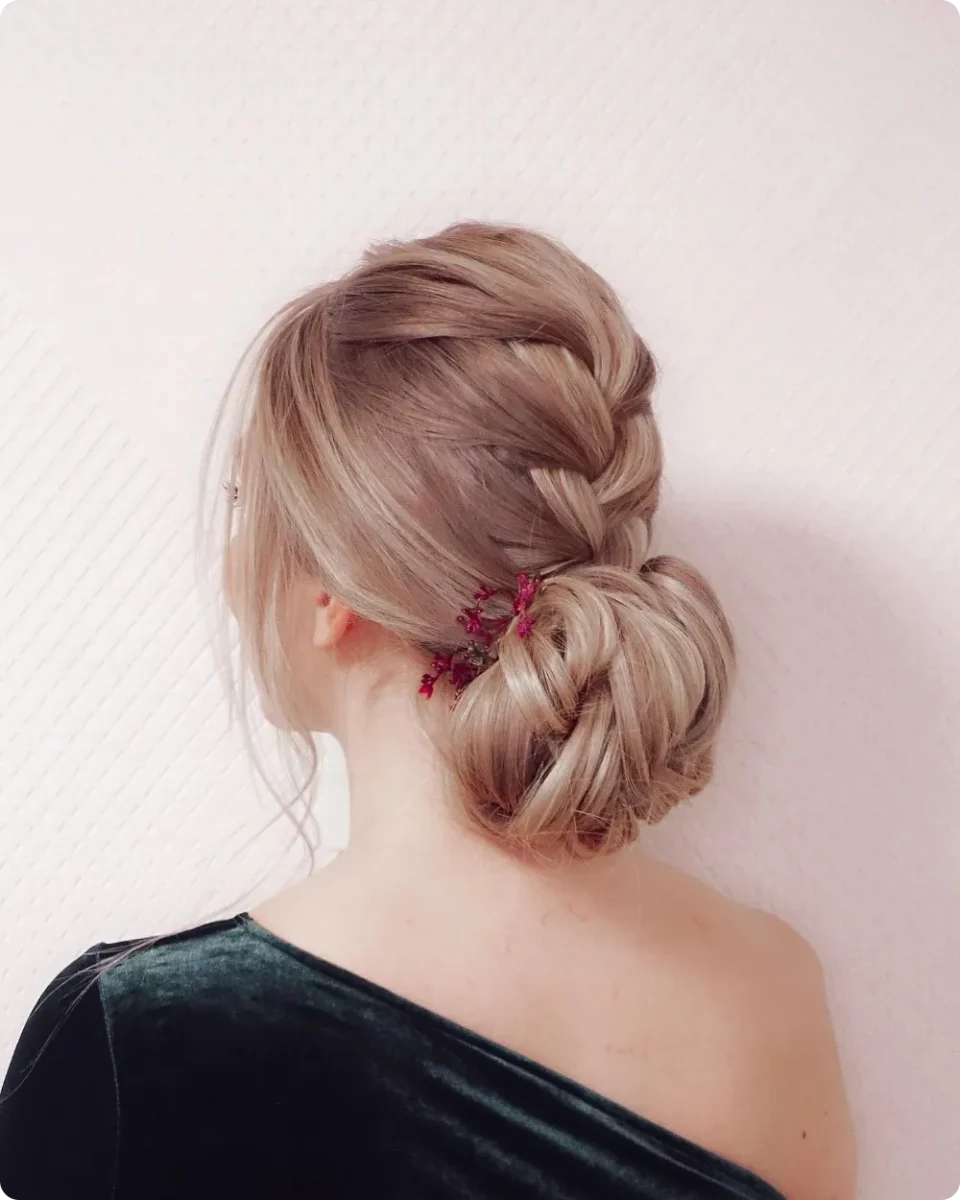Dutch braid leading into a twisted low bun on ash blonde hair with tiny dried magenta flowers tucked at the seam