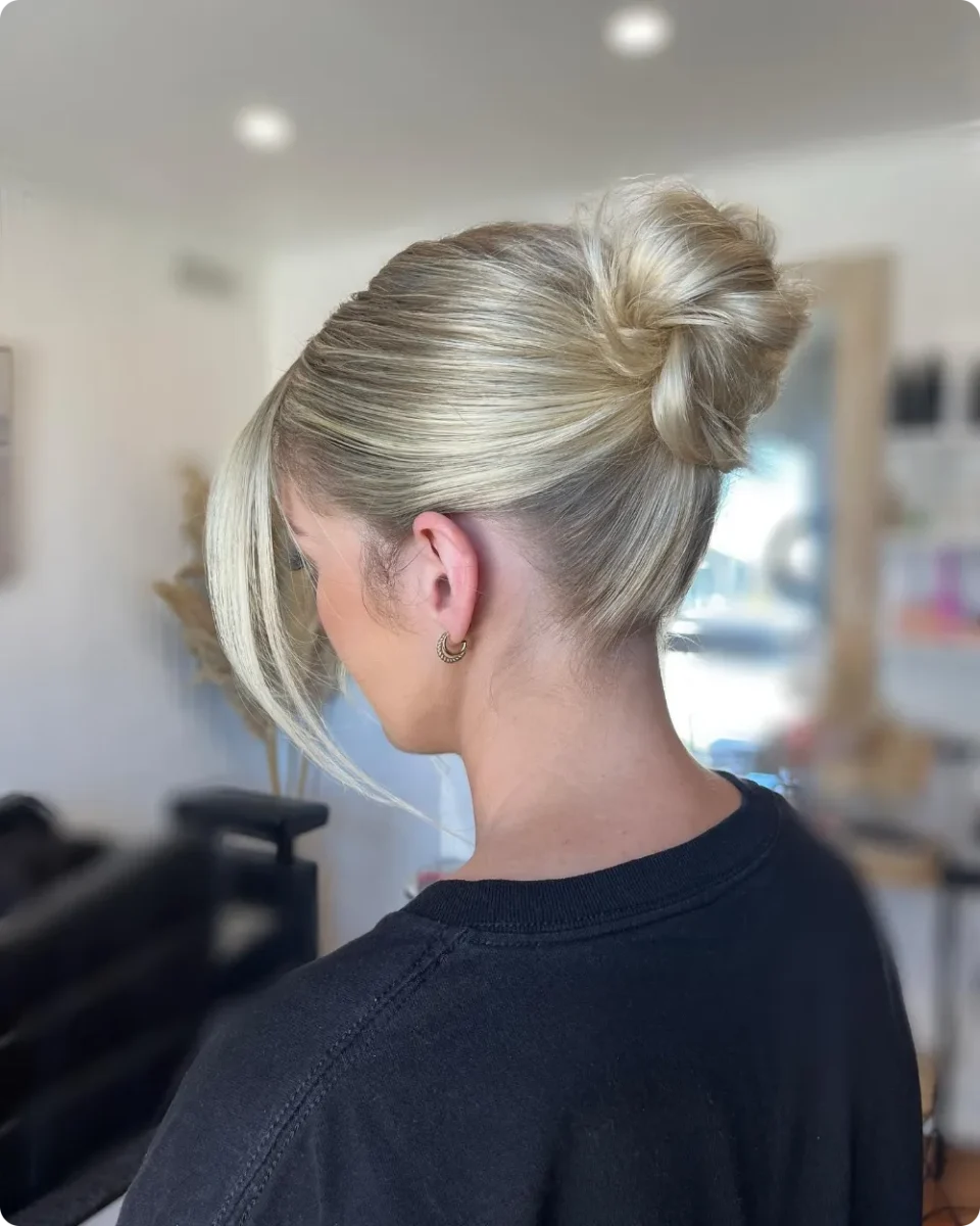 High knotted bun on cool blonde hair with a long front section swept across the cheek and small gold hoops