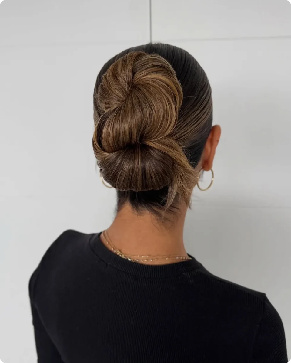 Oversized rolled bun on dark brown hair with caramel highlights, slicked wet look sides, and gold hoop earrings