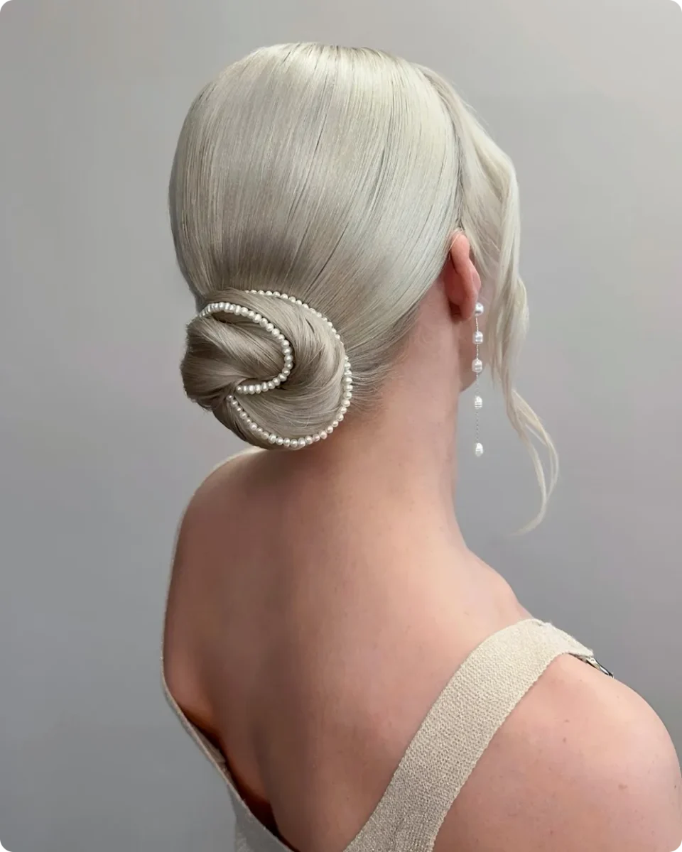 Sleek spiral bun on platinum blonde hair wrapped with a strand of pearls, paired with long pearl drop earrings