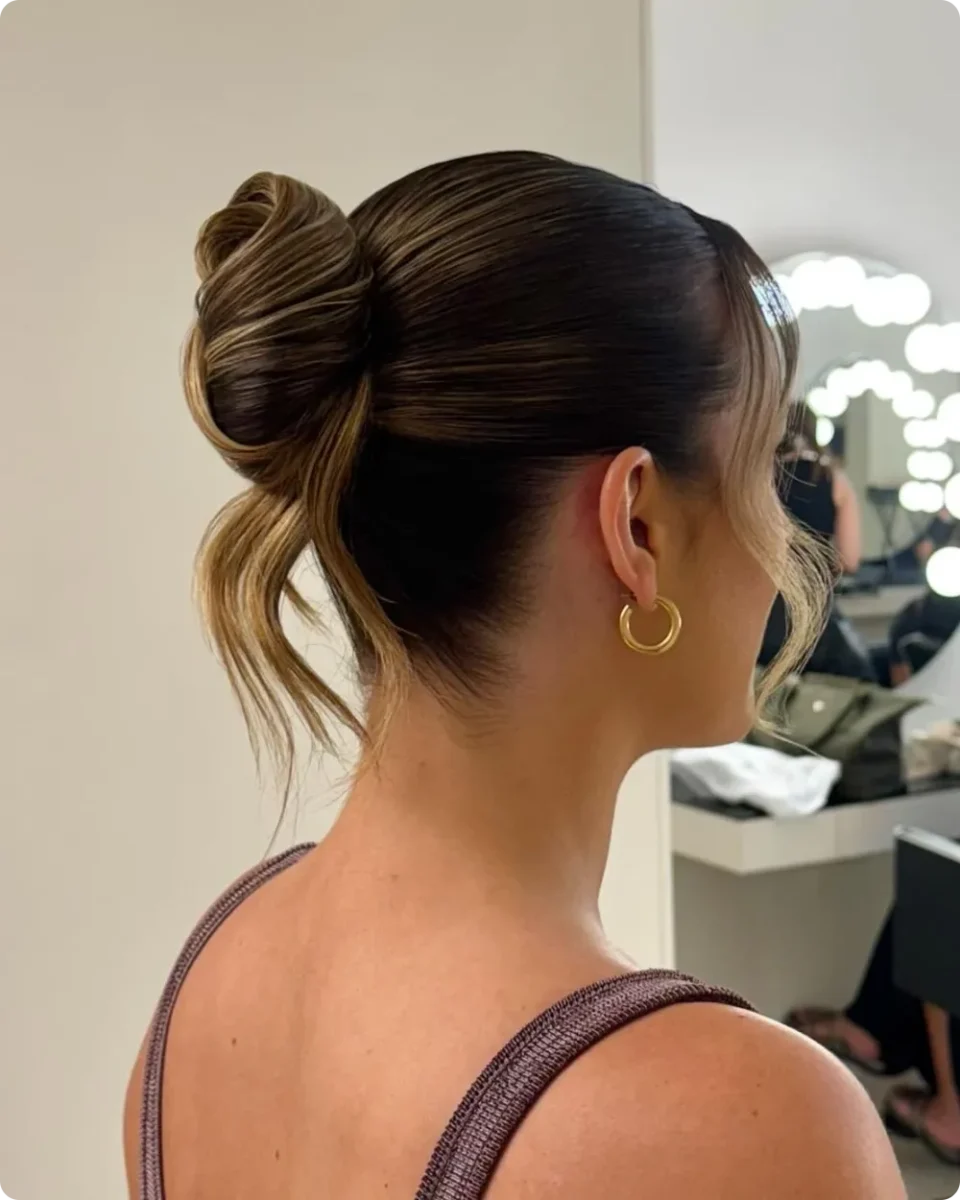 Sleek high bun at the crown on brunette hair with caramel highlights, smooth sides, and a gold hoop earring