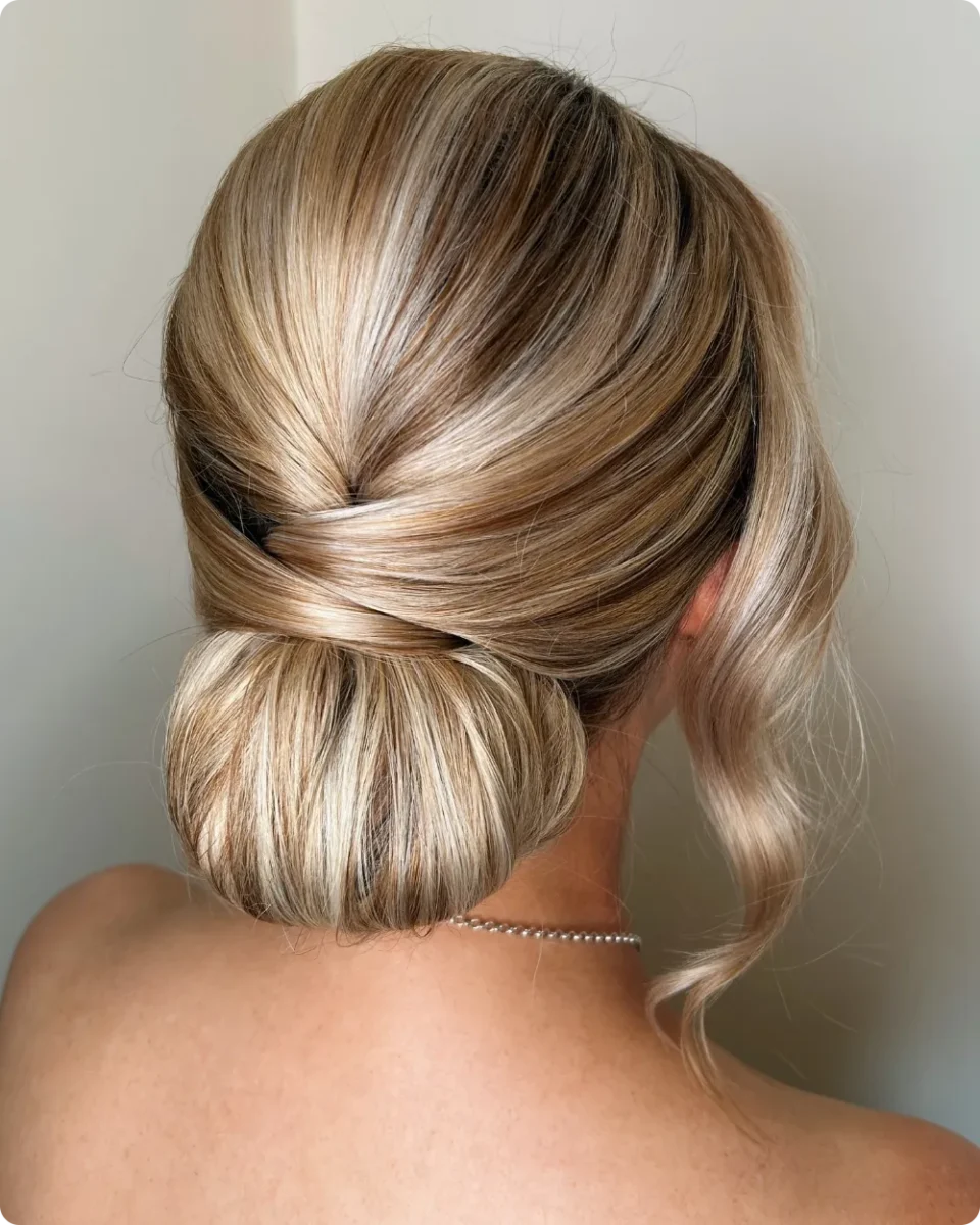 Smooth low bun on blonde long hair with a crossover seam above it and one long curled strand falling at the side
