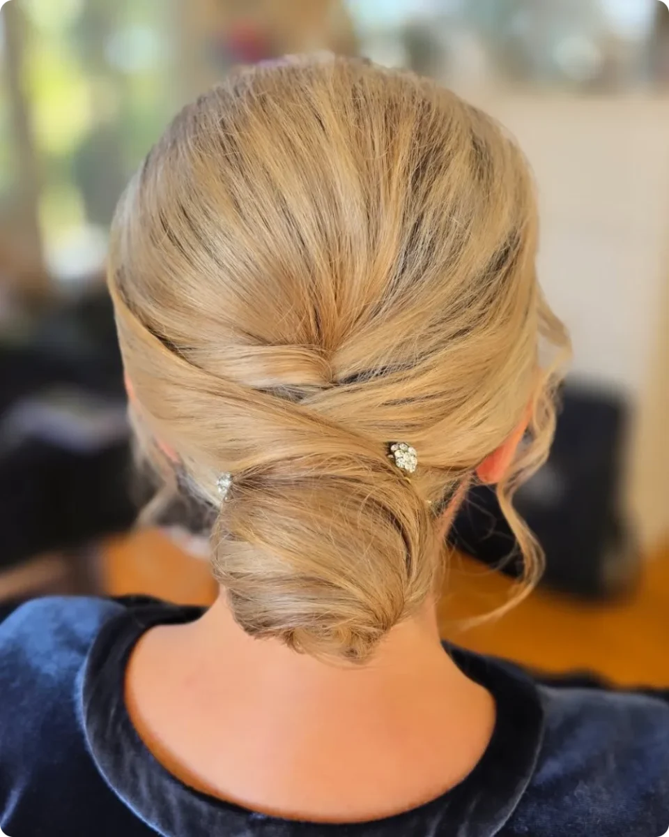 Soft crossover low bun on golden blonde hair with two small crystal pins and loose waves framing the face