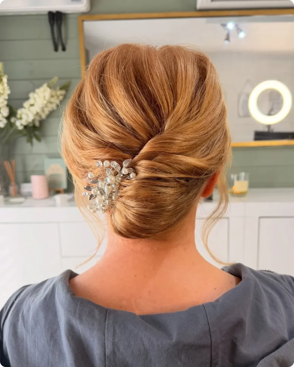 Twisted low chignon on strawberry blonde hair with crown volume and a pearl and leaf hair vine set along the twist