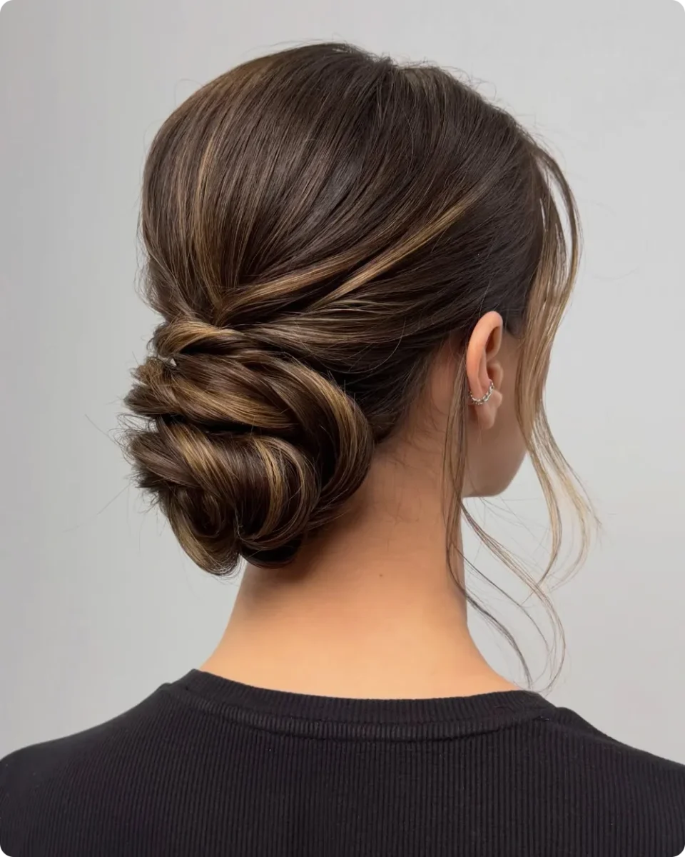 Twisted low bun on brunette hair with caramel ribbons, a deep diagonal side sweep, and long face framing strands