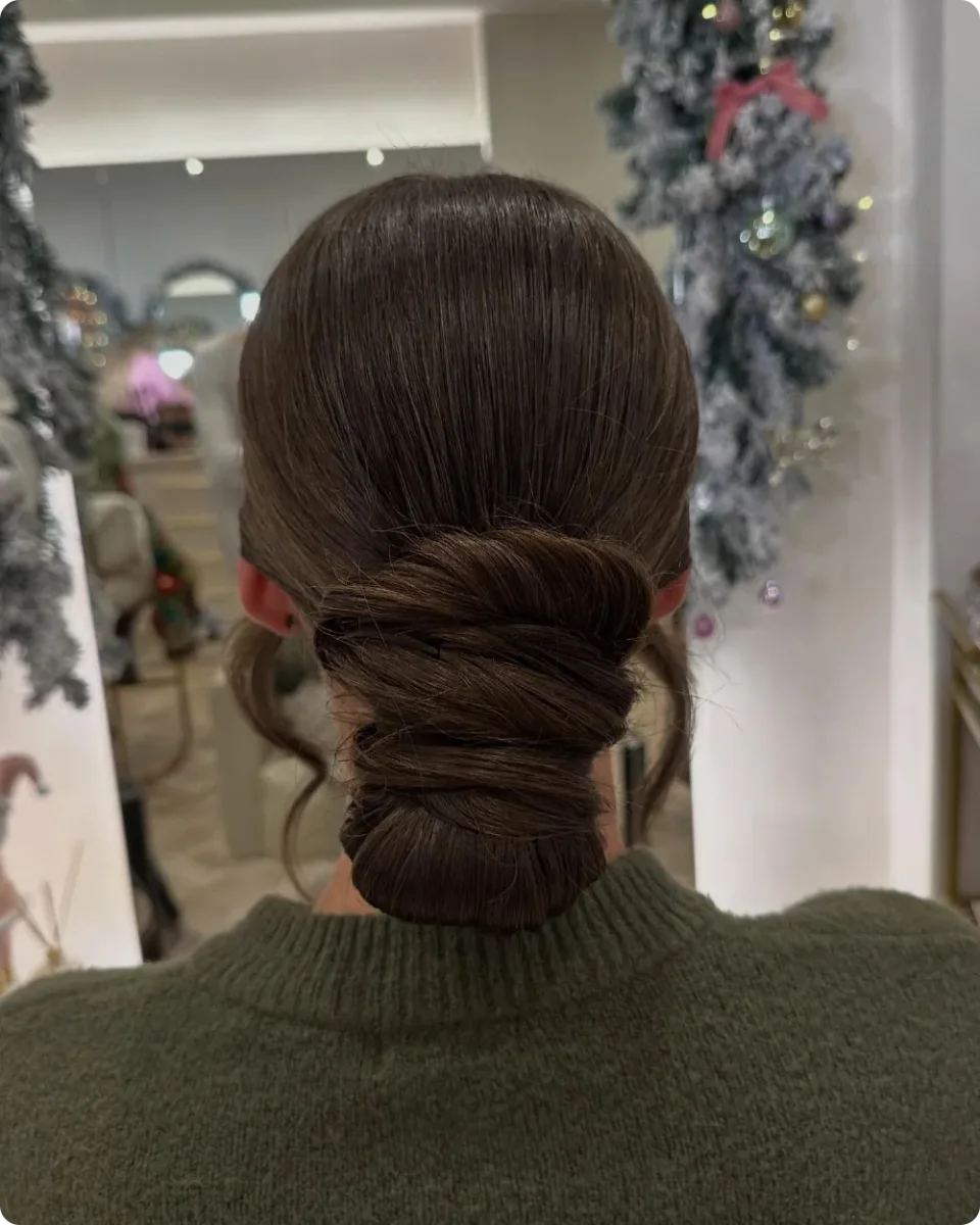 Wrapped low bun on smooth brunette hair with two soft tendrils at the ears, worn with a green knit sweater